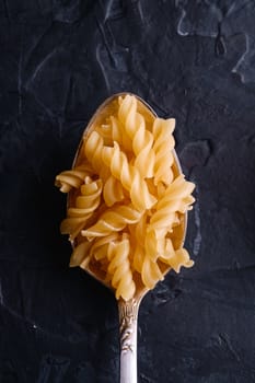 Cutlery spoon with cavatappi uncooked golden wheat curly pasta on dark black textured background, top view