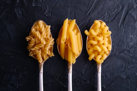 Three cutlery spoons with variety of uncooked golden wheat pasta on dark black textured background, top view