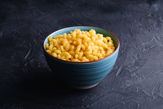 Blue bowl with cavatappi uncooked golden wheat curly pasta on textured dark black background, angle view