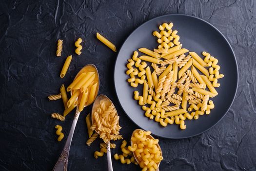 Three cutlery spoons and plate with variety of uncooked golden wheat pasta on dark black textured background, top view