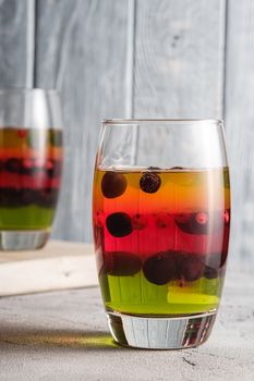 Jelly dessert with berries in glass on old wooden cutting board, sweet colorful layered pudding, stone concrete background, angle view macro