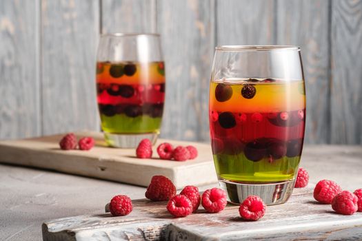 Jelly dessert with berries in glass on old wooden cutting board, sweet colorful layered pudding, stone concrete background, angle view macro