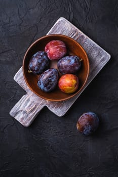Fresh sweet plum fruits in brown wooden bowl on old cutting board, black textured background, top view