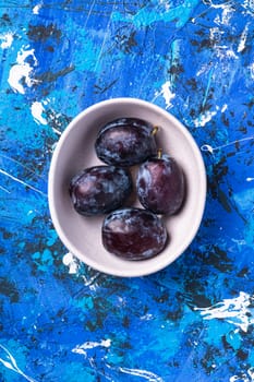 Fresh sweet plum fruits in white bowl on blue abstract background, top view