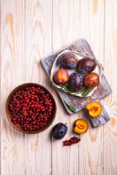 Fresh sweet plum fruits whole and sliced in plate with rosemary leaves on old cutting board with red currant berries in wooden bowl, wood table background, top view