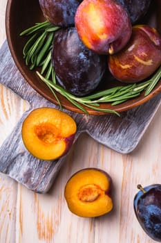 Fresh sweet plum fruits whole and sliced in brown wooden bowl with rosemary leaves on old cutting board, wood table background, top view