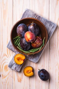 Fresh sweet plum fruits whole and sliced in brown wooden bowl with rosemary leaves on old cutting board, wood table background, top view