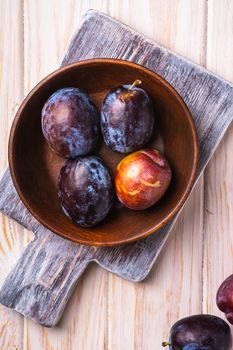 Fresh sweet plum fruits in brown wooden bowl on old cutting board, wood table background, top view