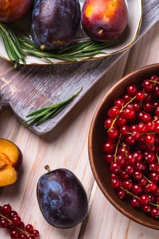 Fresh sweet plum fruits whole and sliced in plate with rosemary leaves on old cutting board with red currant berries in wooden bowl, wood table background, angle view