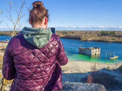 Hiker on mountain top with smartphone. Woman is standing and taking a picture on smartphone. Abandoned Quarry Of Rummu, Estonia. Scenic View Of Land Against Clear Blue Sky. Panoramic View.