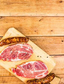 Pork chops with condiments and smoked sausage on a wooden cutting board over wooden table, meat for bbq, top view, copy space, barbeque concept