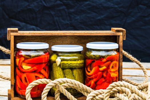 Wooden crate with glass jars with pickled red bell peppers and pickled cucumbers (pickles) isolated. Jars with variety of pickled vegetables. Preserved food concept in a rustic composition.