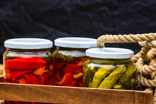 Wooden crate with glass jars with pickled red bell peppers and pickled cucumbers (pickles) isolated. Jars with variety of pickled vegetables. Preserved food concept in a rustic composition.