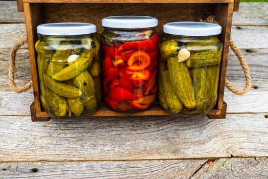 Glass jars with pickled red bell peppers and pickled cucumbers (pickles) isolated in wooden crate. Jars with variety of pickled vegetables. Preserved food concept in a rustic composition.