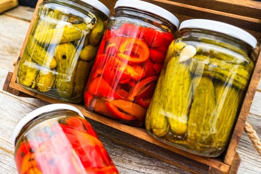 Glass jars with pickled red bell peppers and pickled cucumbers (pickles) isolated in wooden crate. Jars with variety of pickled vegetables. Preserved food concept in a rustic composition.