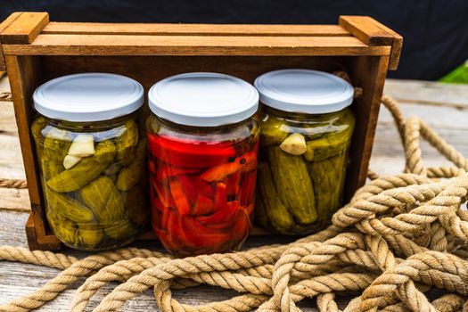 Glass jars with pickled red bell peppers and pickled cucumbers (pickles) isolated in wooden crate. Jars with variety of pickled vegetables. Preserved food concept in a rustic composition.