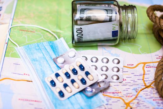 Composition with saving money banknotes in a glass jar with pills and surgical mask on map. Concept of investing and keeping money for healthcare and travel, close up isolated.