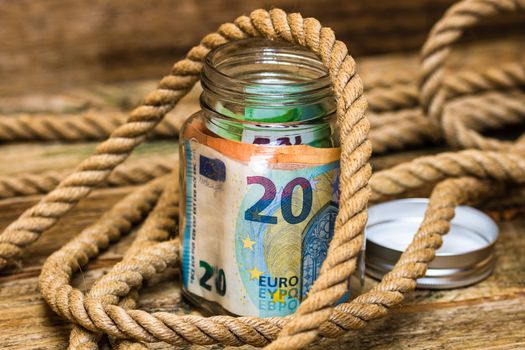 Composition with saving money banknotes in a glass jar surrounded by thick strong rope. Concept of investing and keeping money, close up isolated.
