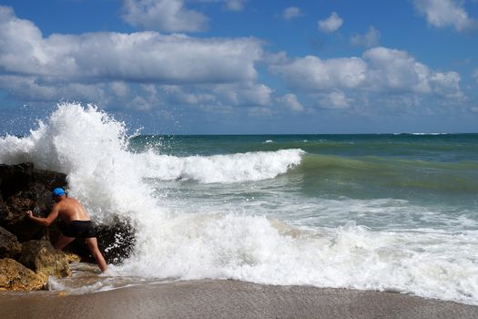 a man holding on to a coastal stone next to a stormy sea wave.