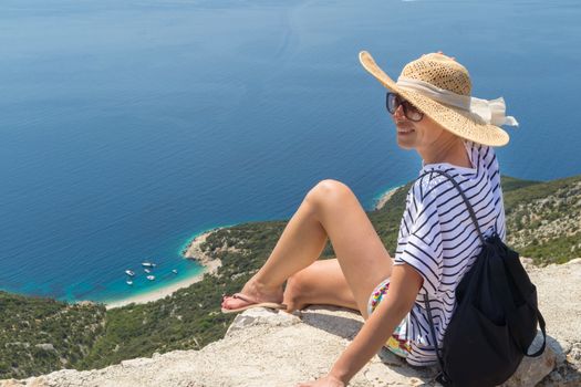 Active sporty woman on summer vacations sitting on old stone wall at Lubenice village, wearing straw hat and beach backpack enjoying beautiful coastal view of Cres island, Croatia.