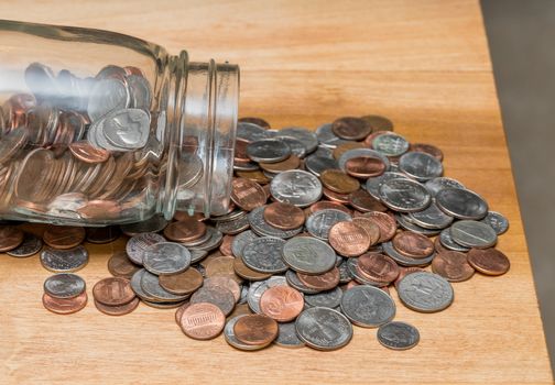 Hundreds of US coins pouring out of savings jar on wooden table as concept for shortage of loose change in USA