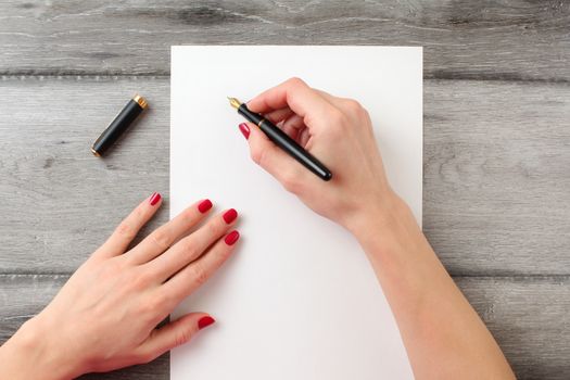 View from above to woman's hands with red nails, holding black fountain ink pen with gold nib, ready to write something on empty piece of paper laying on gray wood table.