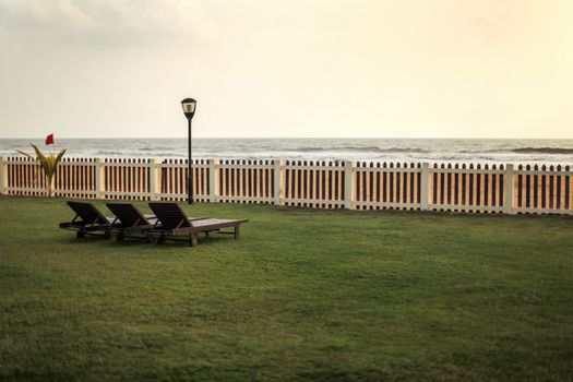 View from hotel resort to empty sunbeds on green lawn, beach and sea behind wooden fence after sunset.