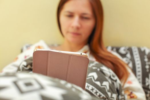 Young woman in pyjama, laying in bed, covered with duvet, working with her tablet before sleeping.