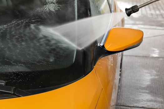 Detail on dark yellow car front window and rear mirror being washed with jet water spray in carwash.