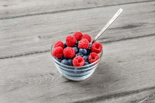 Small glass bowl with spoon full of blueberries and raspberries ready to eat on gray wood table.