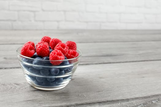 Small glass bowl full of blueberries and raspberries on gray wood table.