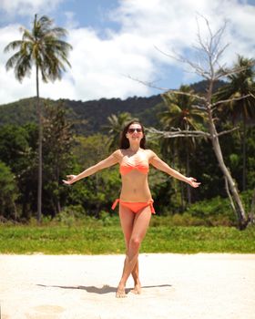 Young woman in red bikini standing on the fine sand beach, arms spread, with palms in background. Klong Jak Beach, Koh Lanta, Thailand