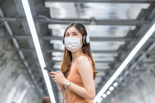 Young Asian woman passenger wearing surgical mask and listening music via smart mobile phone in subway train interchange when traveling in big city at Covid19 outbreak, Infection and Pandemic concept