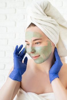 Spa and wellness. Natural cosmetics. Self care. Close up of happy young caucasian woman in white bath towels wearing gloves applying clay face mask looking at the mirror