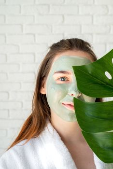 Spa and wellness. Natural cosmetics. Self care. Portrait of a happy young woman with a facial mask holding a monstera leaf