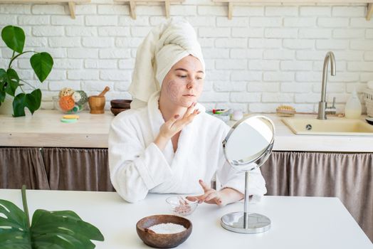 Spa and wellness. Natural cosmetics. Self care. Happy young woman applying face scrub on her face in her home kitchen looking at the mirror