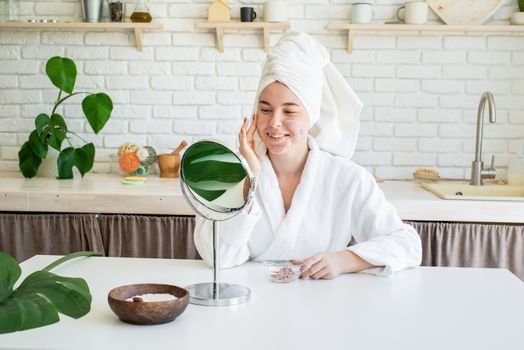 Spa and wellness. Natural cosmetics. Self care. Happy young woman applying face scrub on her face in her home kitchen looking at the mirror