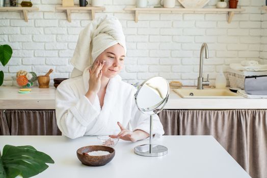 Spa and wellness. Natural cosmetics. Self care. Happy young woman applying face scrub on her face in her home kitchen looking at the mirror