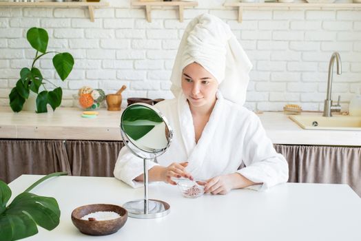 Spa and wellness. Natural cosmetics. Self care. Happy young woman applying face scrub on her face in her home kitchen looking at the mirror