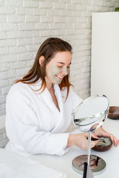 Spa and wellness. Natural cosmetics. Self care. Young smiling caucasian woman wearing bathrobes appplying clay face mask looking at the mirror