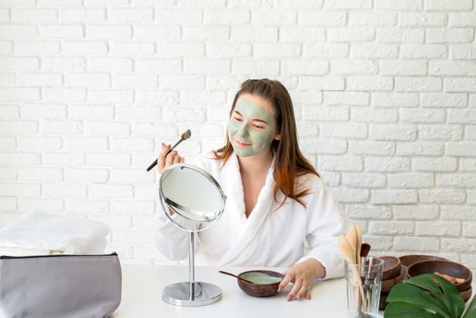 Spa and wellness. Natural cosmetics. Self care. Young smiling caucasian woman wearing bathrobes appplying clay face mask looking at the mirror