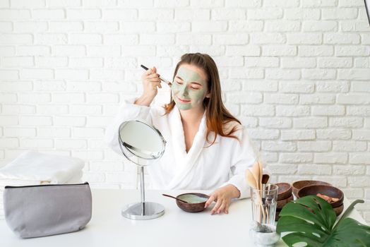 Spa and wellness. Natural cosmetics. Self care. Young smiling caucasian woman wearing bathrobes appplying clay face mask looking at the mirror