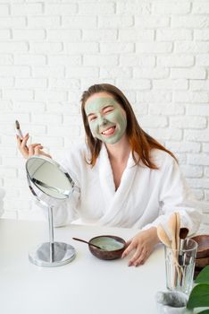 Spa and wellness. Natural cosmetics. Self care. Young smiling caucasian woman wearing bathrobes appplying clay face mask making funny face