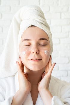 Spa and wellness concept. Selfcare. Natural cosmetics. Young caucasian woman in white bath towels applying face cream doing spa procedures, eyes closed
