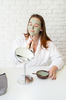 Spa and wellness. Natural cosmetics. Self care. Young smiling caucasian woman wearing bathrobes appplying clay face mask looking at the mirror