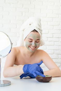 Spa and wellness. Natural cosmetics. Self care. Close up of happy young caucasian woman in white bath towels wearing gloves applying clay face mask looking at the mirror