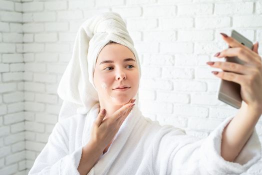 Spa and wellness. Natural cosmetics. Self care. Happy young caucasian woman in white bath towel applying face cream at home doing selfie
