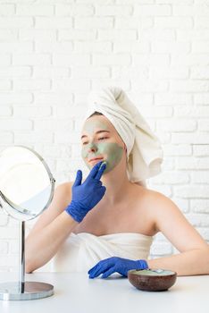 Spa and wellness. Natural cosmetics. Self care. Close up of happy young caucasian woman in white bath towels wearing gloves applying clay face mask looking at the mirror