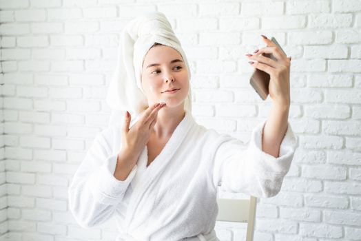 Spa and wellness. Natural cosmetics. Self care. Happy young caucasian woman in white bath towel applying face cream at home doing selfie