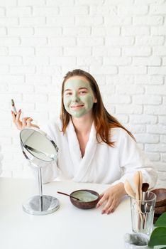 Spa and wellness. Natural cosmetics. Self care. Young smiling caucasian woman wearing bathrobes appplying clay face mask looking at the mirror